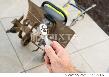 A ratchet tool is being held near a garden tiller resting on a concrete patio. Maintenance work on the garden equipment, focusing on the tool and machine. A ratchet tool is being held near a garden tiller resting on a concrete patio. Maintenance work on the garden equipment, focusing on the tool and machine. 143593070