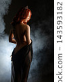 Red haired woman with crown and tattooed back draped in black sheer fabric, standing in smoke with dramatic backlighting 143593182