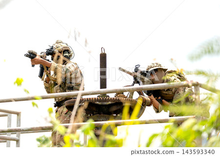 Two soldiers in camouflage gear aiming rifles from elevated metal platform amid bright foliage and overexposed sky Two soldiers in camouflage gear aiming rifles from elevated metal platform amid bright foliage and overexposed sky 143593340