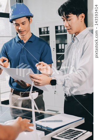 Two Asian engineers wearing hardhats and safety vests discussing solar panel installation and renewable energy project in the office. 143593414