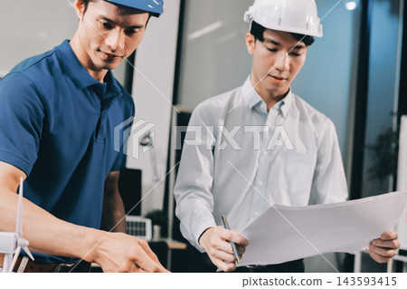 Two Asian engineers wearing hardhats and safety vests discussing solar panel installation and renewable energy project in the office. Two Asian engineers wearing hardhats and safety vests discussing solar panel installation and renewable energy project in the office. 143593415