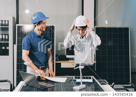 Two Asian engineers wearing hardhats and safety vests discussing solar panel installation and renewable energy project in the office. Two Asian engineers wearing hardhats and safety vests discussing solar panel installation and renewable energy project in the office. 143593416