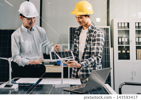 Two Asian engineers wearing hardhats and safety vests discussing solar panel installation and renewable energy project in the office. Two Asian engineers wearing hardhats and safety vests discussing solar panel installation and renewable energy project in the office. 143593417