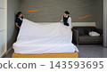 Two smiling professional women hotel housekeepers in uniform working together to spread clean white sheet over bed in  modern guest room 143593695