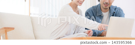 Banner, a Japanese couple in their 40s, sits on the sofa in their brightly lit living room, searching for information together on a laptop. Banner, a Japanese couple in their 40s, sits on the sofa in their brightly lit living room, searching for information together on a laptop. 143595366