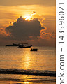 Captivating golden hour sunset over tranquil ocean waters. Sun rays pierce through clouds, illuminating a silhouetted fishing boat and a person. A tropical island adds to this serene seascape. 143596021
