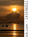 Captivating golden sunset over a tropical sea, featuring the sun reflecting on the calm water. Silhouettes of a boat, a person wading, and a distant island create a serene scene. 143596022