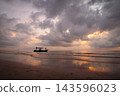 Golden hour glow illuminates a dramatic cloudy sky over a tranquil tropical beach. A solitary fishing boat with people is silhouetted against the vibrant reflections on the wet sand and ocean. 143596023