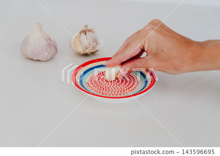 Hand grinds a peeled clove on a ceramic garlic grater plate next to tomatoes and whole bulbs on a table. Kitchen tool for mincing garlic and ginger in home cooking and healthy meal preparation. Hand grinds a peeled clove on a ceramic garlic grater plate next to tomatoes and whole bulbs on a table. Kitchen tool for mincing garlic and ginger in home cooking and healthy meal preparation. 143596695