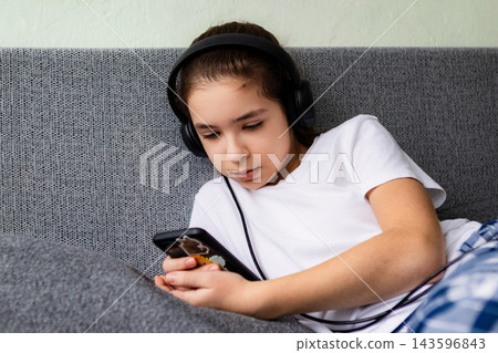 Preteen girl listening on headphones while relaxing with smartphone on a gray couch at home Preteen girl listening on headphones while relaxing with smartphone on a gray couch at home 143596843