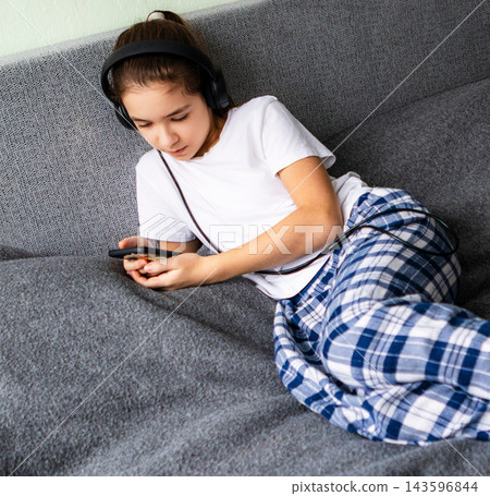 Girl wearing headphones and pajamas lounging on sofa while using smartphone and listening to music Girl wearing headphones and pajamas lounging on sofa while using smartphone and listening to music 143596844
