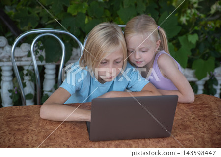 Young boy and girl sharing a laptop, focusing on the screen while engaging in a shared educational or entertainment activity on a patio with a leafy green background Young boy and girl sharing a laptop, focusing on the screen while engaging in a shared educational or entertainment activity on a patio with a leafy green background 143598447