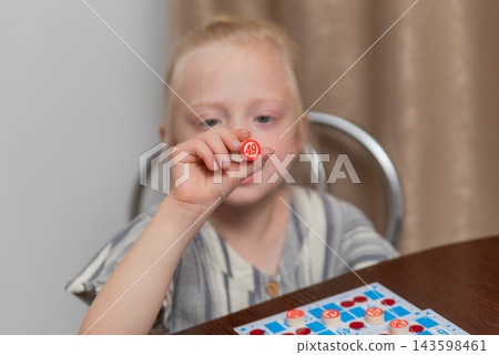 Child concentrating on the game, holding a red number token with 49 on it, participating in a fun family activity with bingo cards visible on the table Child concentrating on the game, holding a red number token with 49 on it, participating in a fun family activity with bingo cards visible on the table 143598461