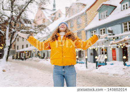 Smiling woman in a bright jacket is having fun with the snow on a snowy city street. Smiling woman in a bright jacket is having fun with the snow on a snowy city street. 143602014