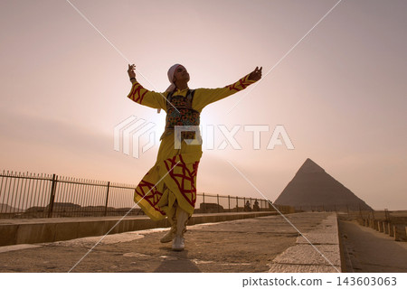 Traditional dancer in vibrant yellow embroidered robe spinning arms outstretched at iconic Giza Pyramids against glowing hazy sunset sky ideal for Traditional dancer in vibrant yellow embroidered robe spinning arms outstretched at iconic Giza Pyramids against glowing hazy sunset sky ideal for 143603063