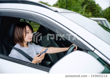 一位年輕女子坐在汽車駕駛座上,看著她的智慧型手機。 一位年輕女子坐在汽車駕駛座上,看著她的智慧型手機。 143603218