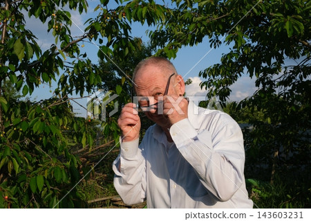 Senior man removing and putting on glasses while smiling at camera near apple tree in sunny garden showing confidence and playful charm Senior man removing and putting on glasses while smiling at camera near apple tree in sunny garden showing confidence and playful charm 143603231
