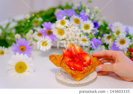 Female hand holding flaky strawberry pastry with blooming white and purple flowers creating a fresh summer dessert scene close up Female hand holding flaky strawberry pastry with blooming white and purple flowers creating a fresh summer dessert scene close up 143603335