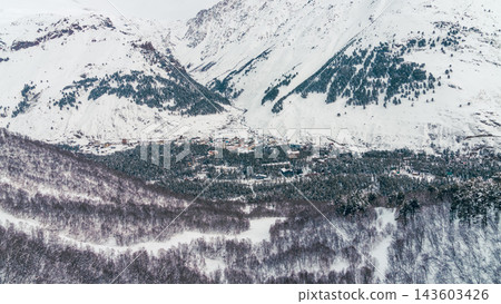 Aerial winter panorama of mountain village nestled beneath steep snow-covered peaks surrounded by dense forest and frozen clear Aerial winter panorama of mountain village nestled beneath steep snow-covered peaks surrounded by dense forest and frozen clear 143603426