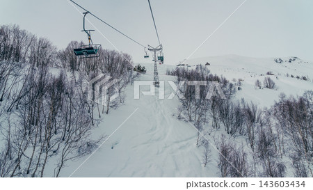 Empty ski lift chairs suspended above snow-covered slope with birch trees and distant snowy ridge Empty ski lift chairs suspended above snow-covered slope with birch trees and distant snowy ridge 143603434