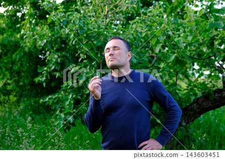 Middle aged country man holding grass stem in teeth and standing thoughtfully near apple tree in rural garden Middle aged country man holding grass stem in teeth and standing thoughtfully near apple tree in rural garden 143603451
