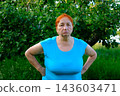 Senior red haired woman standing with hands on hips looking tired sad and disappointed beneath apple tree in summer garden 143603471