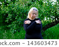 Middle aged blonde woman embracing herself and looking down with sadness while reflecting on aging in garden 143603473
