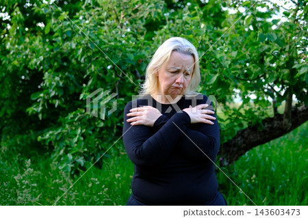 Middle aged blonde woman embracing herself and looking down with sadness while reflecting on aging in garden Middle aged blonde woman embracing herself and looking down with sadness while reflecting on aging in garden 143603473
