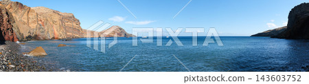 Panoramic view of a rocky volcanic bay with clear blue ocean, rugged cliffs and anchored sailboats under a bright sky in Madeira, Portugal. 143603752