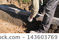Construction workers spread wet concrete beside freshly set curb edging on sunlit soil, using gloves and hand tools to shape foundation edge at outdoor building site with close ground level view. 143603769