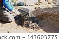 Construction worker kneels beside foundation trench, spreading and smoothing fresh concrete with trowel to form straight edge for residential house project in bright daylight outdoors. 143603775