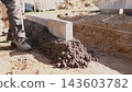 Worker leveling fresh concrete beside straight concrete edge in groundwork for house construction, with soil trench, guide string, boots, sunlight, and outdoor building site details. 143603782