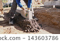 Construction worker sets concrete curb block on prepared foundation at residential building site, using leveling string, soil trench, and fresh mortar in bright daylight. 143603786