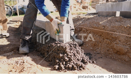 Construction worker sets concrete curb block on prepared foundation at residential building site, using leveling string, soil trench, and fresh mortar in bright daylight. 143603786