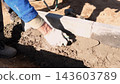 Construction worker wearing gloves levels wet mortar between concrete blocks in outdoor foundation trench, using hand trowel on dirt ground under bright sunlight and strong shadows. 143603789