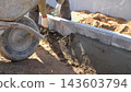 Close view of hands shaping wet concrete along curb edge at residential building site, with wheelbarrow, soil mound, and bright daylight highlighting masonry progress and careful finishing work. 143603794