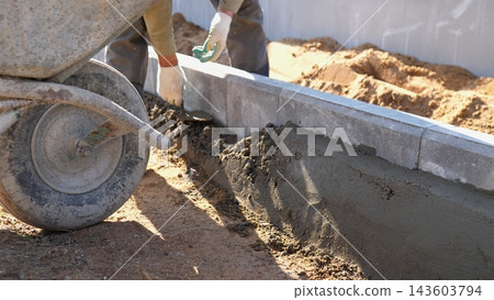 Close view of hands shaping wet concrete along curb edge at residential building site, with wheelbarrow, soil mound, and bright daylight highlighting masonry progress and careful finishing work. Close view of hands shaping wet concrete along curb edge at residential building site, with wheelbarrow, soil mound, and bright daylight highlighting masonry progress and careful finishing work. 143603794