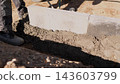 Construction worker boots beside fresh concrete and mortar around light gray block wall section in trench, showing groundwork, masonry, and outdoor building progress under strong sunlight. 143603799