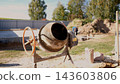 Old cement mixer on sunny construction site beside fresh concrete foundation, sand, tools, and worker in background, showing home building work under clear daytime light. 143603806