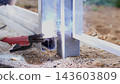 Close view of worker welding metal fencing outdoors, with bright arc light, flying sparks, smoke, protective gloves, and steel parts being joined on gravel ground in daylight 143603809