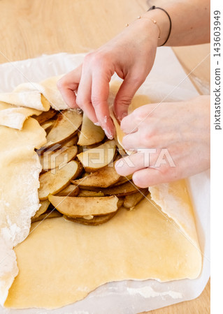 Fruit pastry. Cooking galette. Female baker hand covering sliced pear with flattered dough flour sheet on home kitchen paper parchment. Fruit pastry. Cooking galette. Female baker hand covering sliced pear with flattered dough flour sheet on home kitchen paper parchment. 143603949