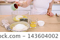 Cooking dough. Treat recipe. Woman chef hand pouring liquid raw egg in bowl with sugar making cupcake batter on home kitchen table. 143604002