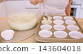 Preparing muffin. Treat recipe. Woman chef hand pouring batter from glass bowl spoon in paper liners in baking tin on home kitchen counter. 143604018