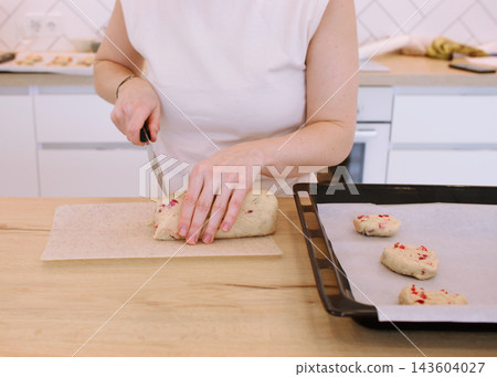 Cookies recipe. Baker work. Woman hand cut biscotti roll log dough with cranberry on home kitchen table placing treat on oven tray. Cookies recipe. Baker work. Woman hand cut biscotti roll log dough with cranberry on home kitchen table placing treat on oven tray. 143604027