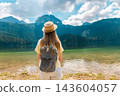 Woman with Backpack Facing Durmitor Mountains Across Black Lake 143604057