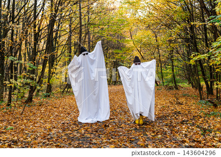 Two people dressed in Halloween ghost costumes stand or walk through an autumn forest surrounded by colorful foliage. The costumes are made from white fabric sheets covering the figures Two people dressed in Halloween ghost costumes stand or walk through an autumn forest surrounded by colorful foliage. The costumes are made from white fabric sheets covering the figures 143604296