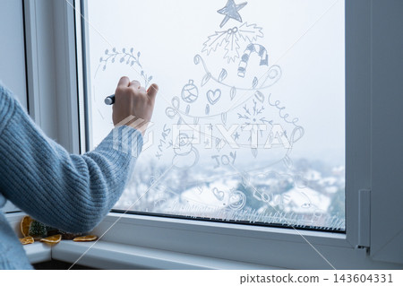 Sustainable responsible holiday decor. Female hand painting Christmas tree painted on window glass indoor at home. Christmas winter decoration. White pattern. Snow art tracery on season. Diy Sustainable responsible holiday decor. Female hand painting Christmas tree painted on window glass indoor at home. Christmas winter decoration. White pattern. Snow art tracery on season. Diy 143604331
