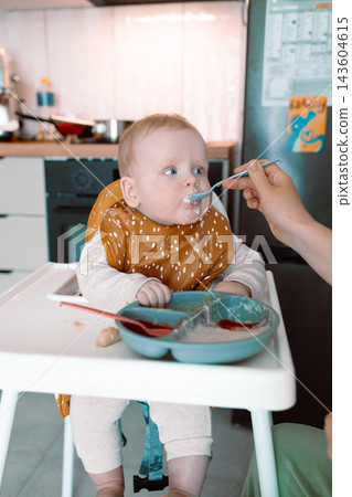 Caring mother hand feeding cute baby boy with spoon in high chair during mealtime at kitchen Caring mother hand feeding cute baby boy with spoon in high chair during mealtime at kitchen 143604615