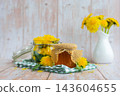Transparent glass of jar dandelion jelly with dandelion flowers in vase. Soft natural lighting highlights the amber color glossy texture and artisanal character of the preserve. Concept of homemade 143604655