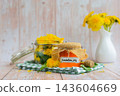Transparent glass of jar DANDELION JELLY text with dandelion flowers in vase. Soft natural lighting highlights the amber color glossy texture and artisanal character of the preserve. Concept of 143604669